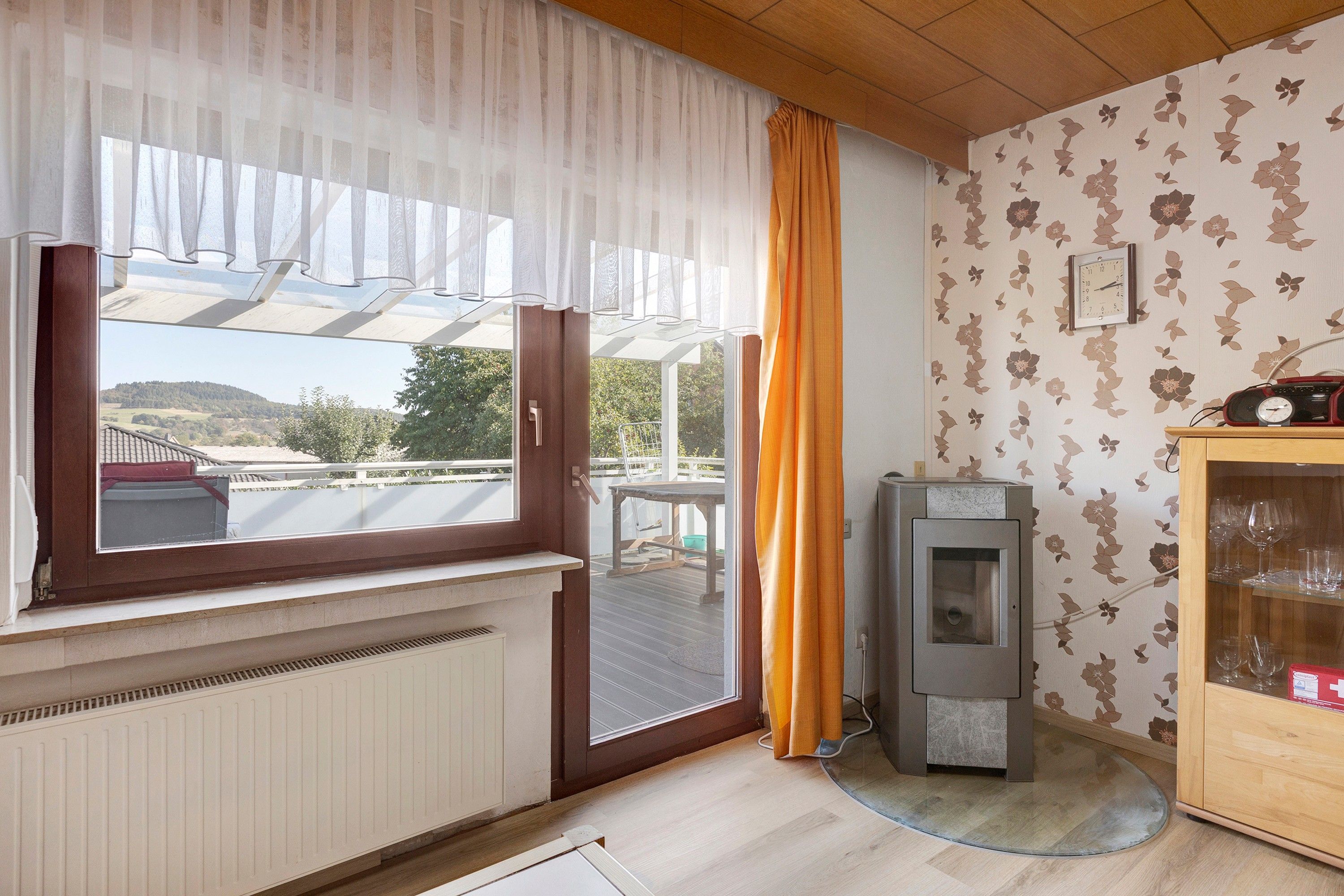 Ein Zimmer mit großem Fenster, Heizkörper, Holzschrank und Wanduhr.