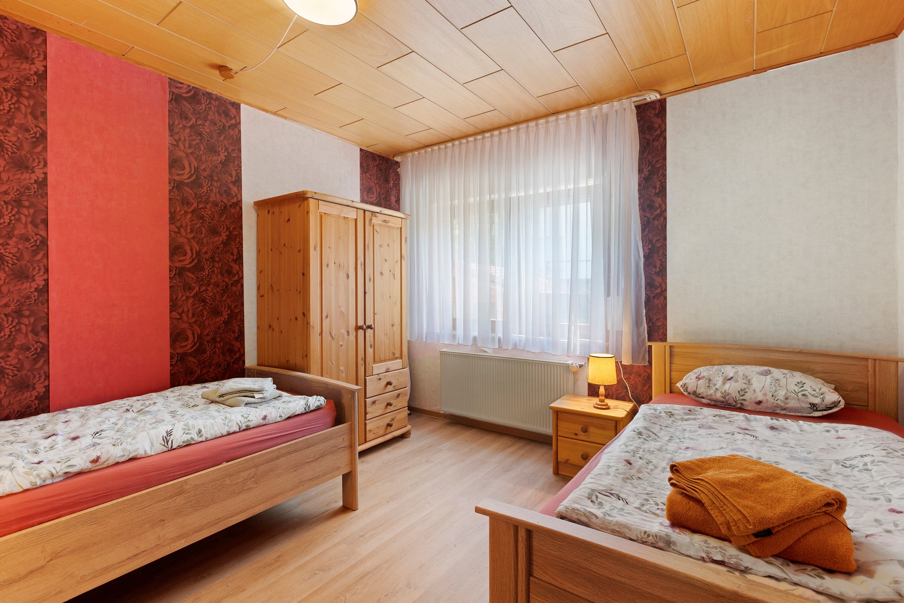 A bedroom with two single beds, a wooden wardrobe, a nightstand with a lamp, and a window with sheer curtains.