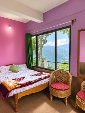 Un dormitorio con una cama, dos sillas de mimbre, una ventana con vistas a montañas y una pared morada.