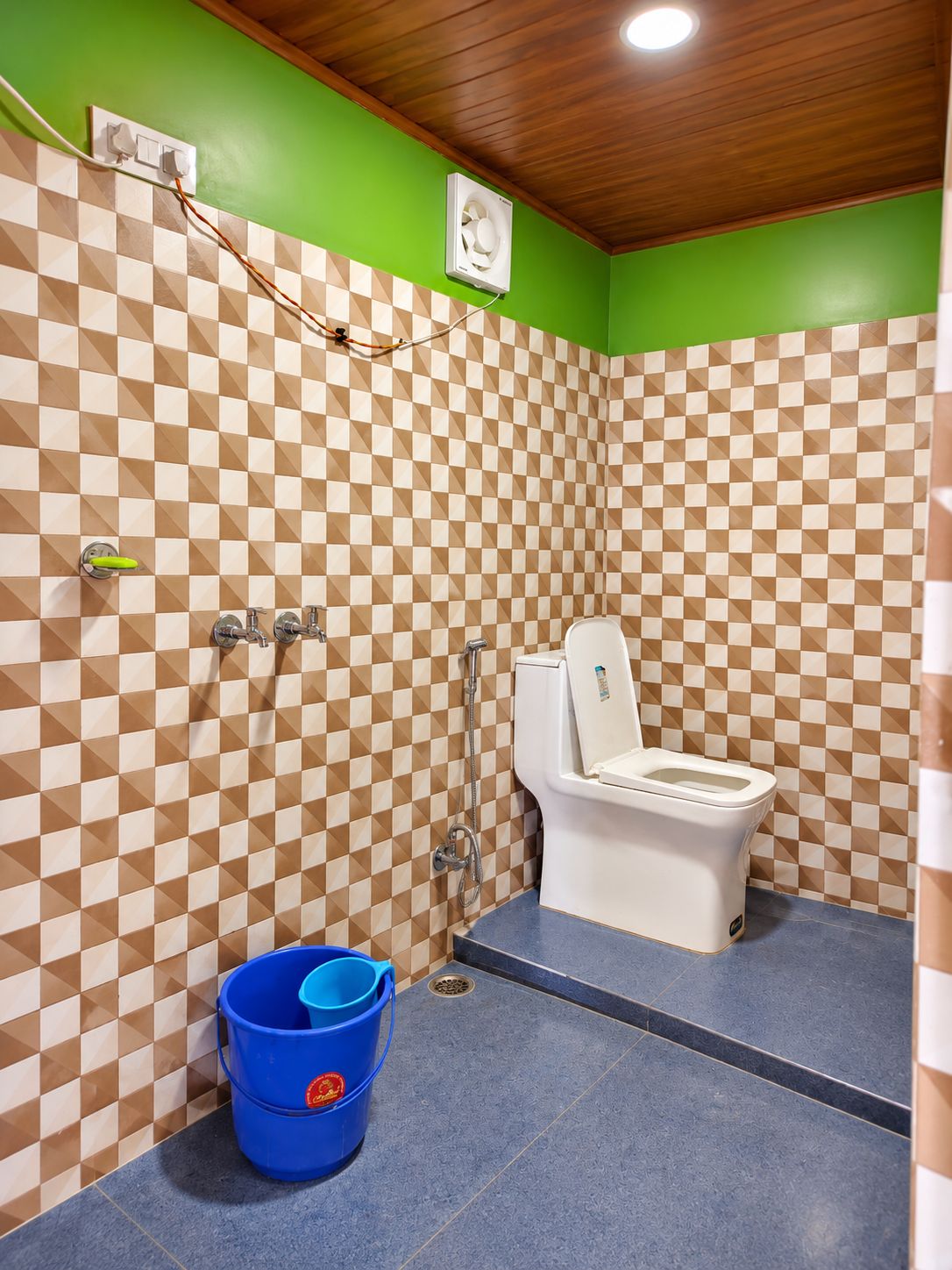 A bathroom with a checkered tile wall, a toilet, a shower, and a blue bucket.
