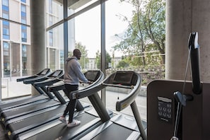 Una persona usando una cinta de correr en un gimnasio con ventanas amplias y vistas a edificios y árboles.