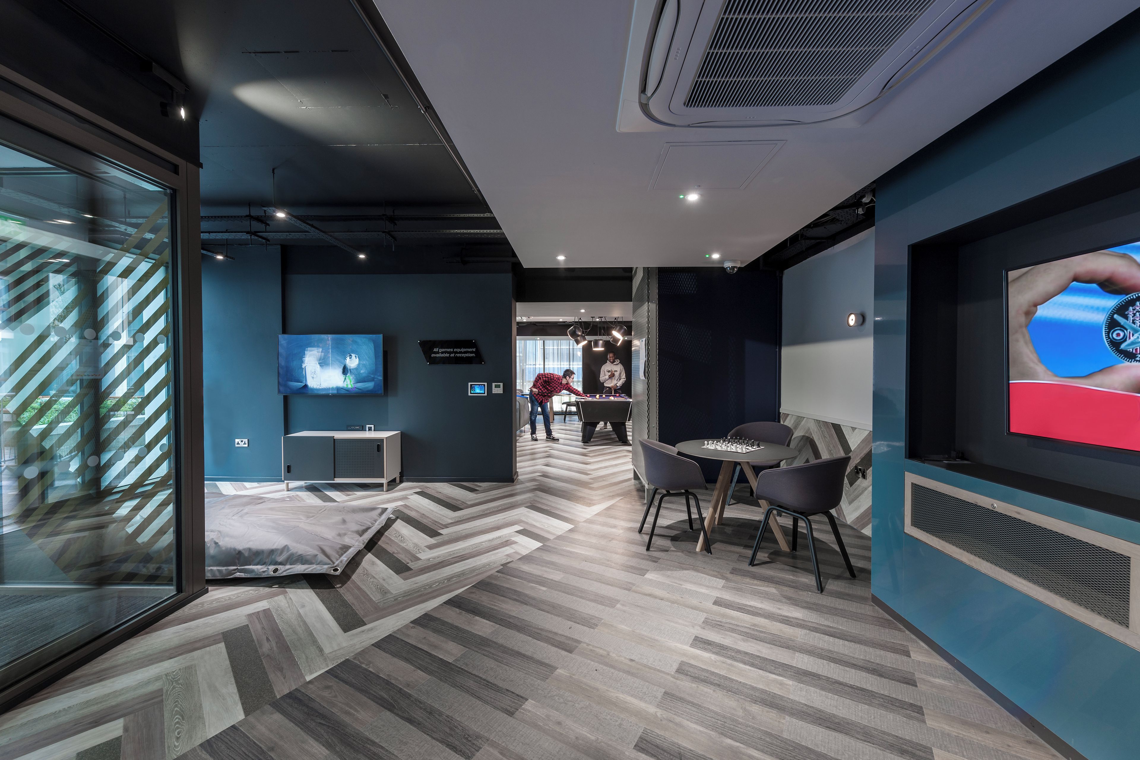 Un área de lounge moderna con piso de patrón espiga, televisor de pantalla plana, mesa de futbolín y un espacio de descanso con sillas.