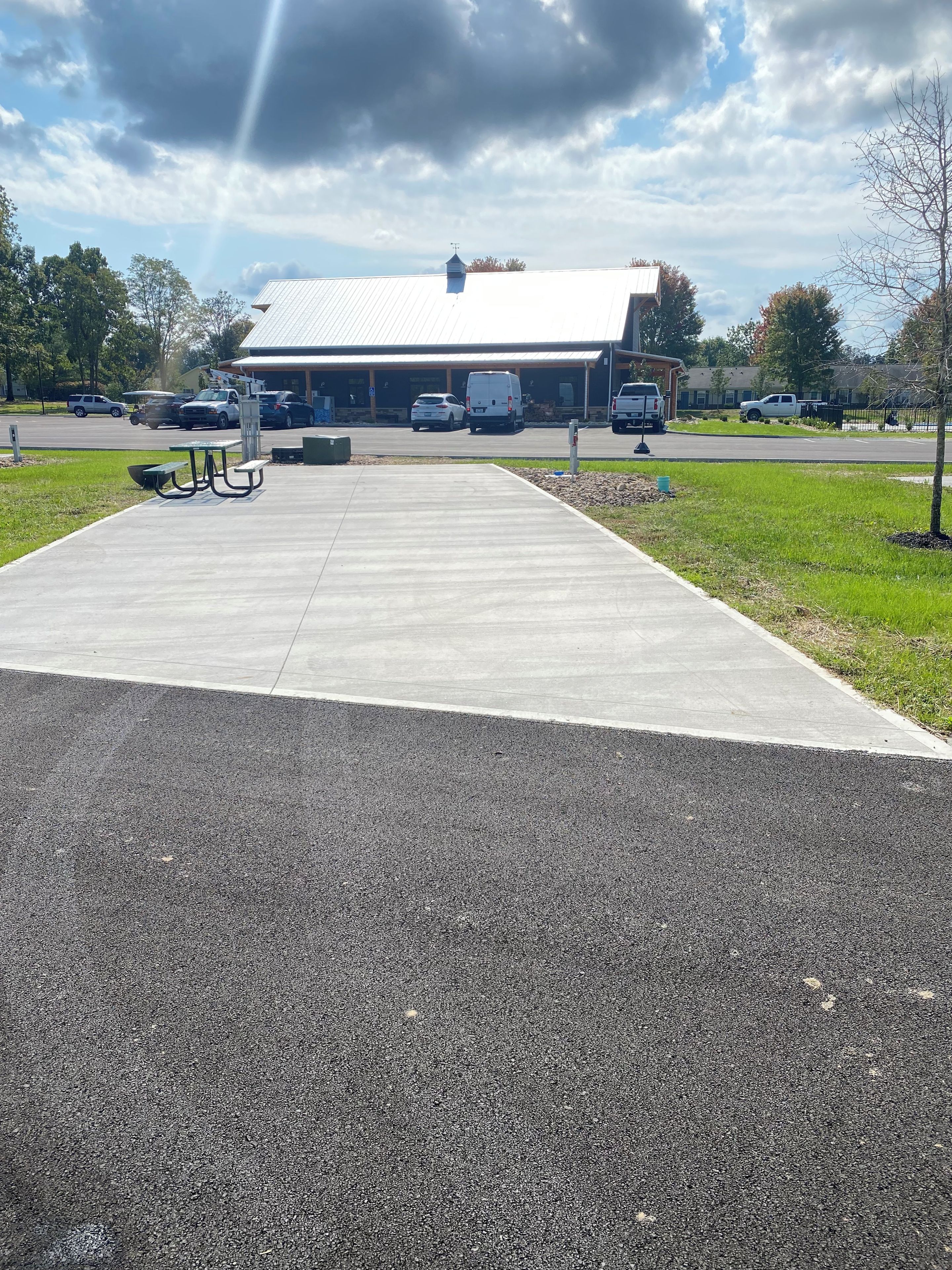Ein gepflasterter Weg führt zu einem Gebäude mit flachem Dach, Picknicktischen und geparkten Fahrzeugen.