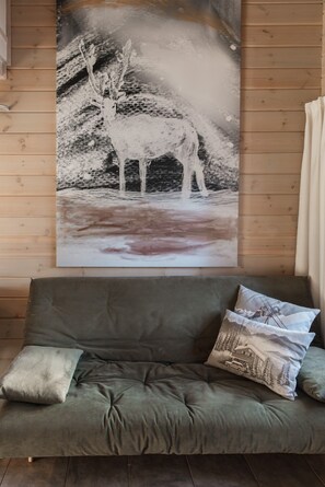 Ein Zimmer mit einem großen Hirschbild an einer Holzwand, einem grauen Sofa mit Kissen und einem Fenster mit Vorhängen.