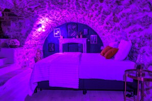 A bedroom with a stone wall backdrop, a bed with a white comforter, and a nightstand with a lamp. - "Le Secret" Suite - Luxury & Private Jacuzzi (Lorgues)