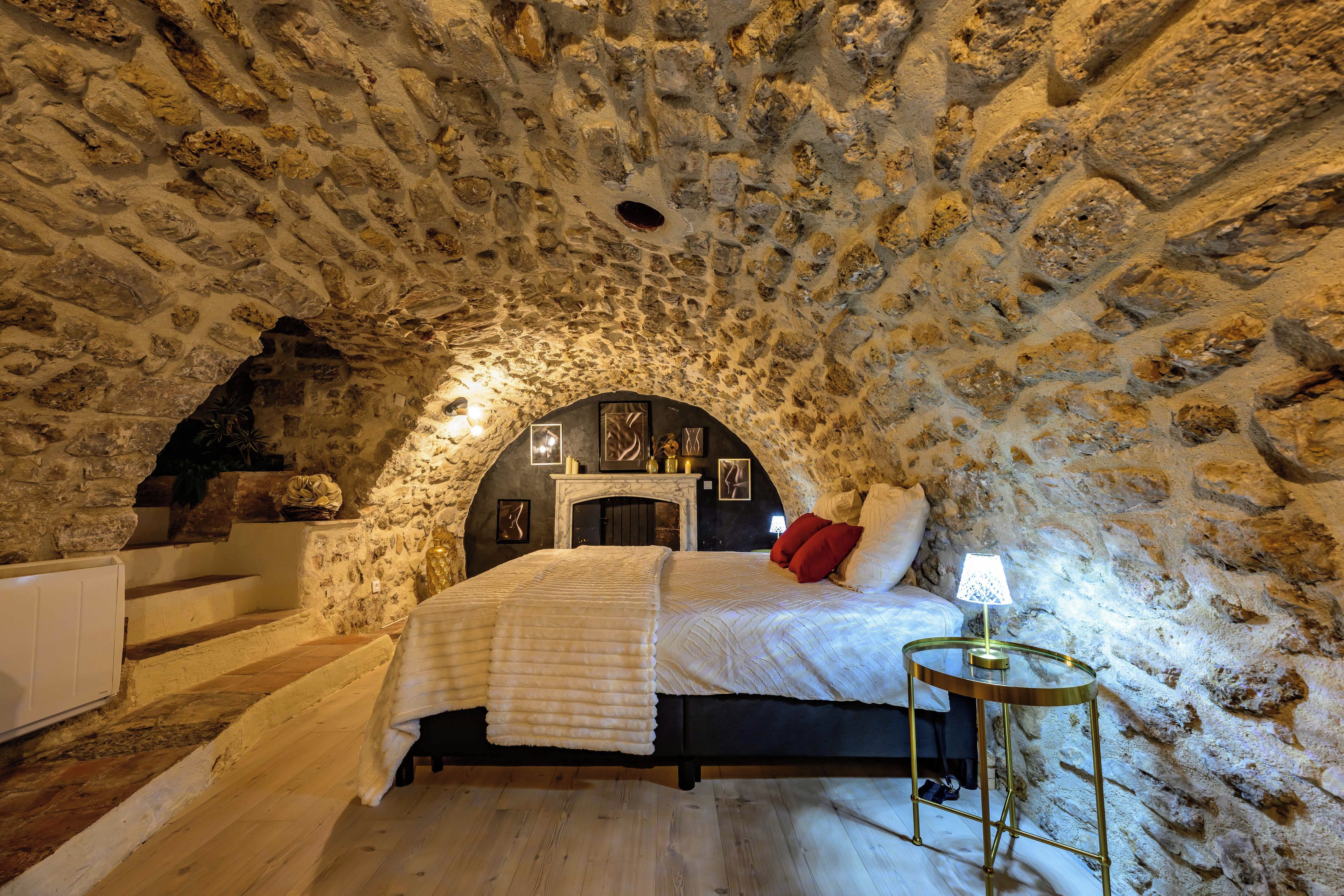 Un dormitorio con techo de piedra, una cama con ropa de cama blanca, una mesita con una lámpara y una chimenea.