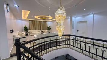A multi-level interior with a grand chandelier, a staircase with black railings, and a seating area with sofas.