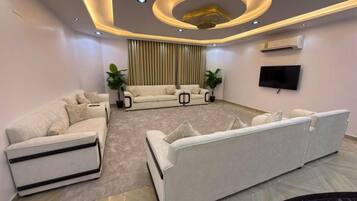 Un salon moderne avec un canapé d’angle, un téléviseur à écran plat fixé au mur et un luminaire décoratif au plafond.