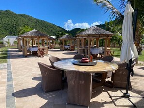 Ein Außenbereich mit einer Terrasse, ausgestattet mit runden Tischen und Korbstühlen, umgeben von hölzernen Pavillons und Palmen.