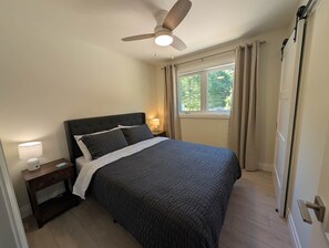 A bedroom with a bed, nightstand, lamp, ceiling fan, window with curtains, and a door.
