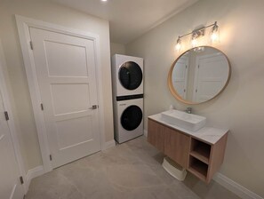 A bathroom with a white door, a wall-mounted washing machine, a sink with a wooden vanity, and a round mirror with lights.