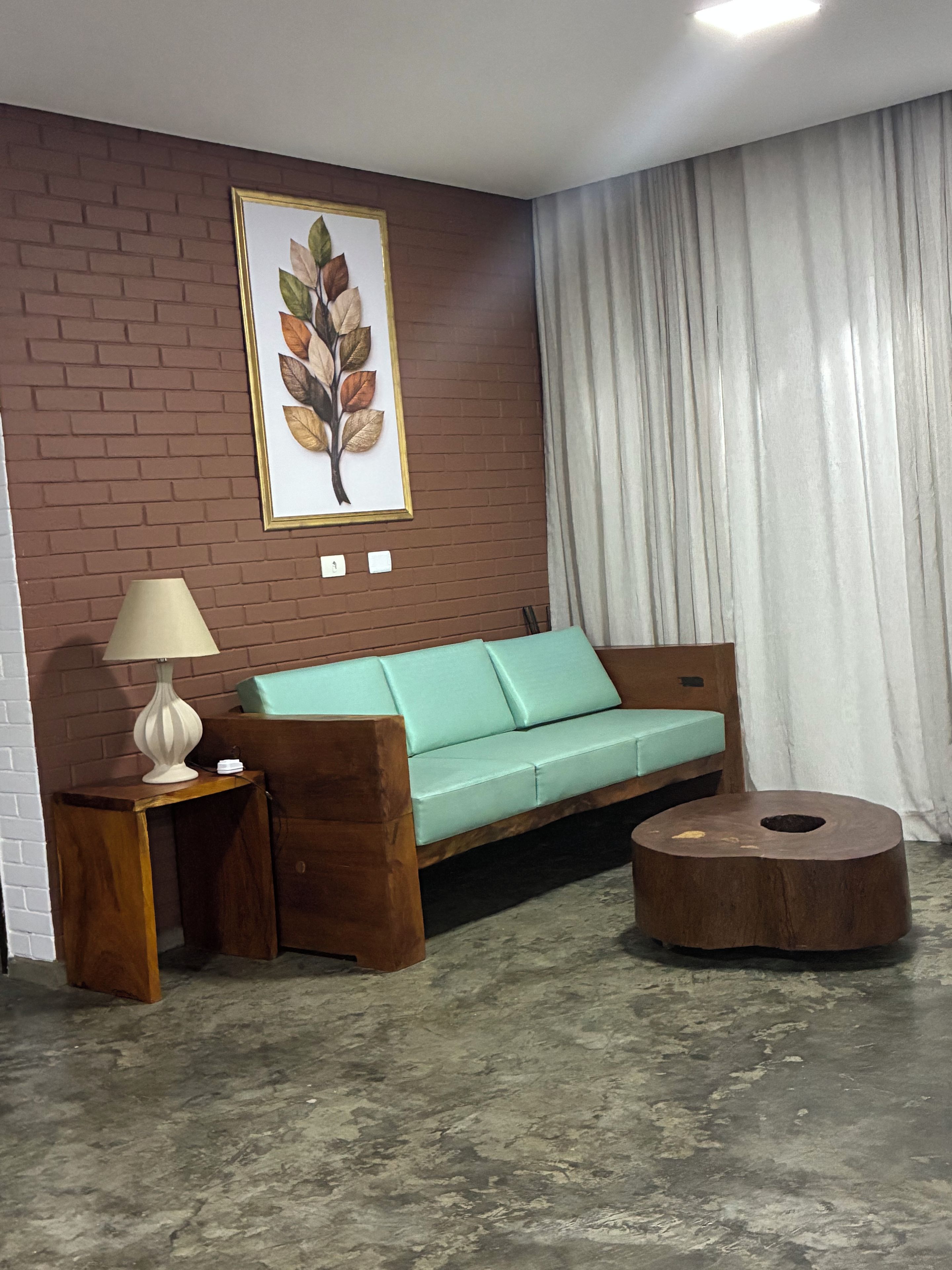 Una habitación con pared de ladrillo, un sofá verde, una mesita de madera con una lámpara, un taburete de madera redondo y una obra de arte con una hoja enmarcada.