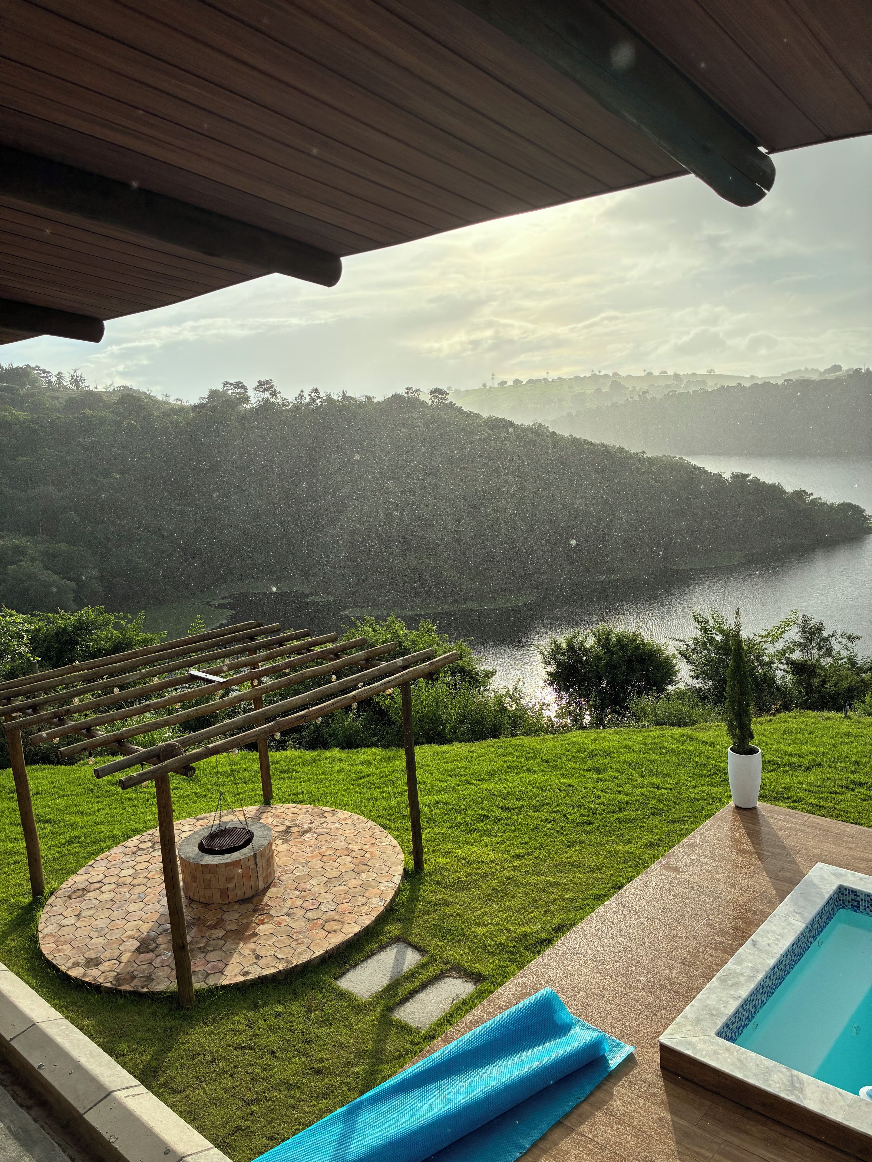 Un área de piscina con una pérgola, un pequeño hogar para fuego y vistas a un lago rodeado de colinas.
