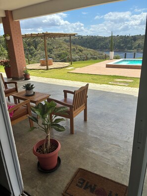 A patio with a wooden table and chairs, potted plants, and a view of a pool and hills. - Casa Pedra do Cavalo | Swimming pool and Paraguaçu River | Pier and boat ramp (Governador Mangabeira)