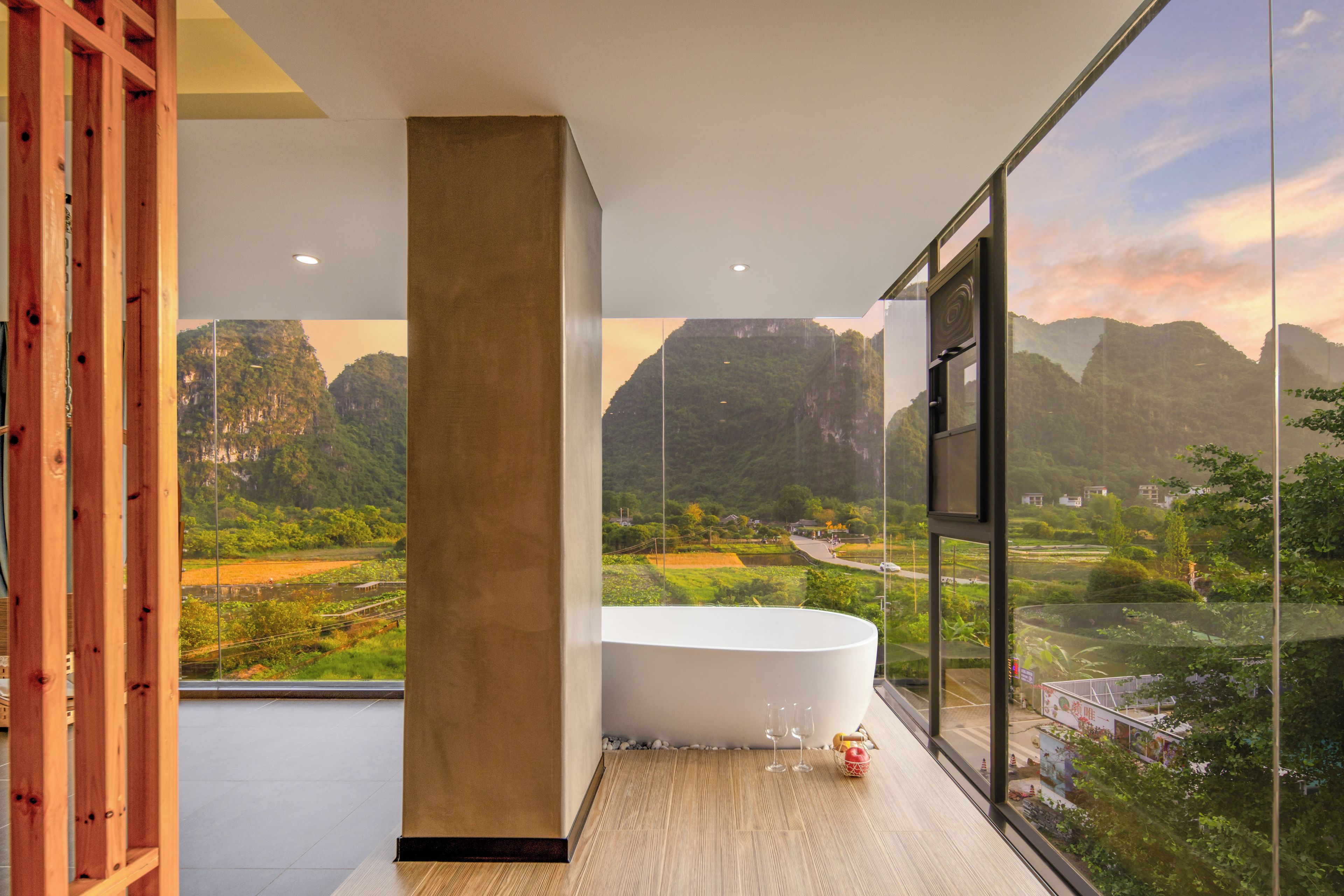 A modern bathroom with a large bathtub, a wooden column, and a view of a mountainous landscape through large windows.