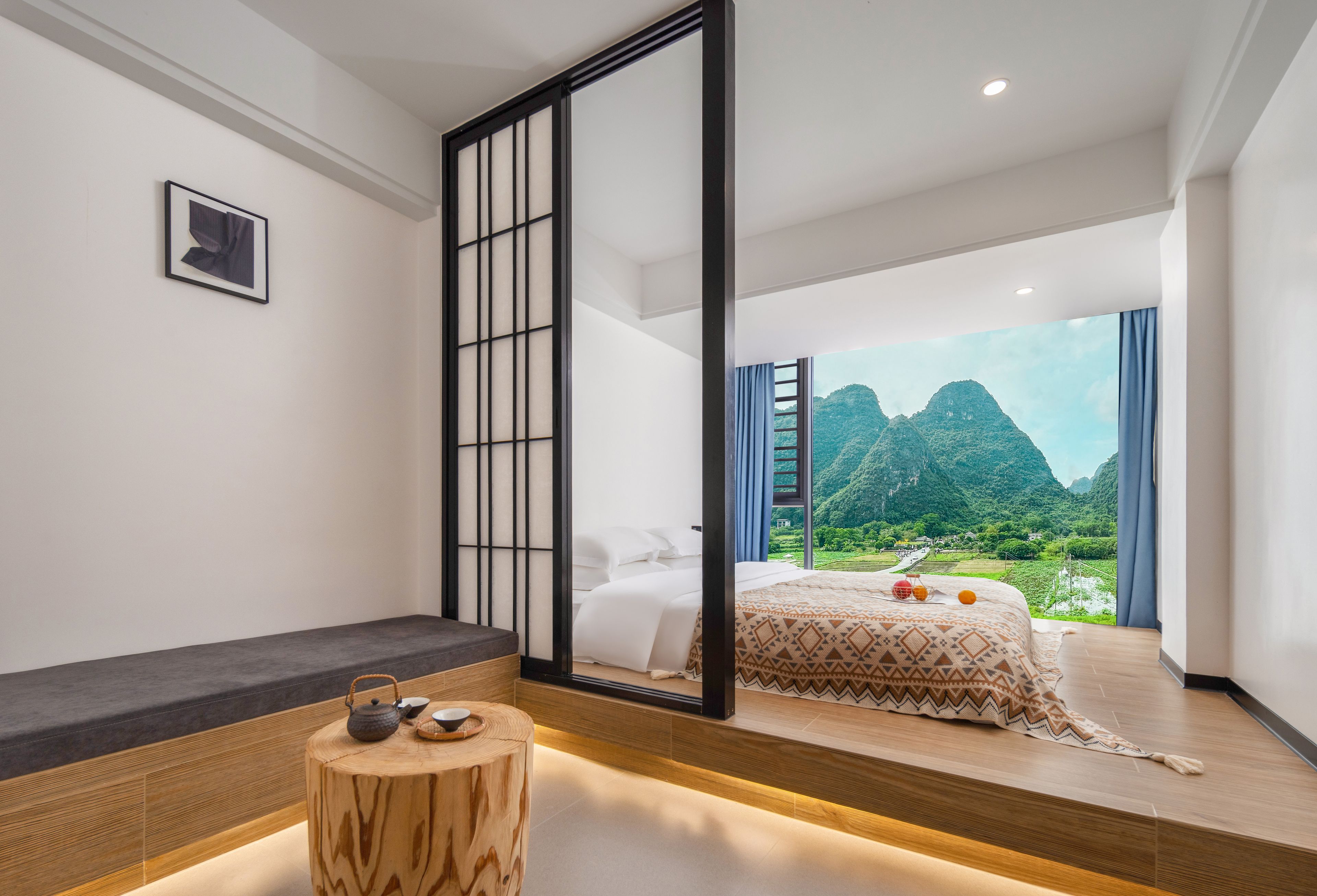 A modern bedroom with a large bed, a wooden stool with a teapot, and a view of a mountainous landscape.