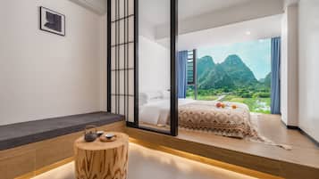 A modern bedroom with a large bed, a wooden stool with a teapot, and a view of a mountainous landscape.
