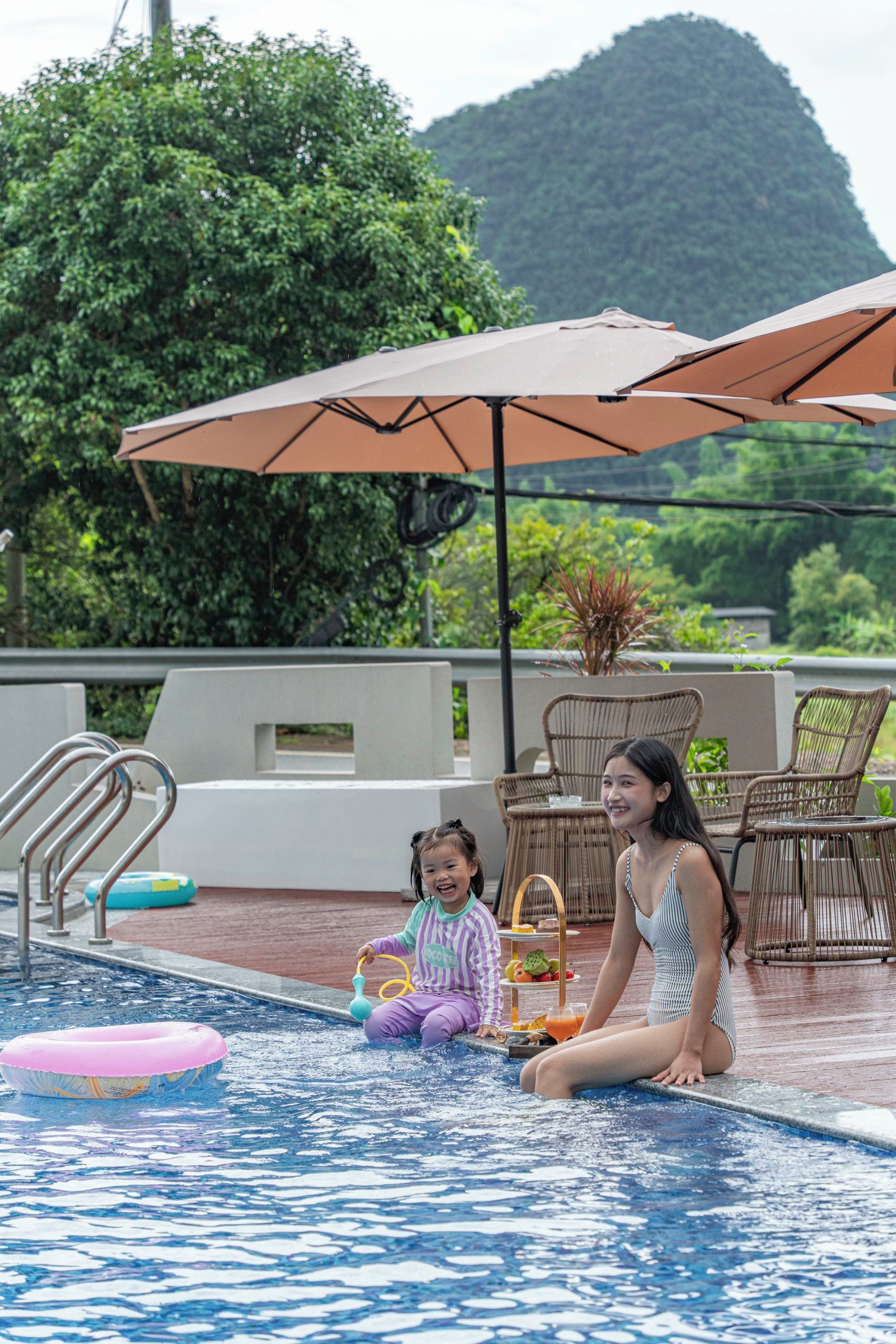 A pool area with a child and an adult, outdoor furniture, and umbrellas.