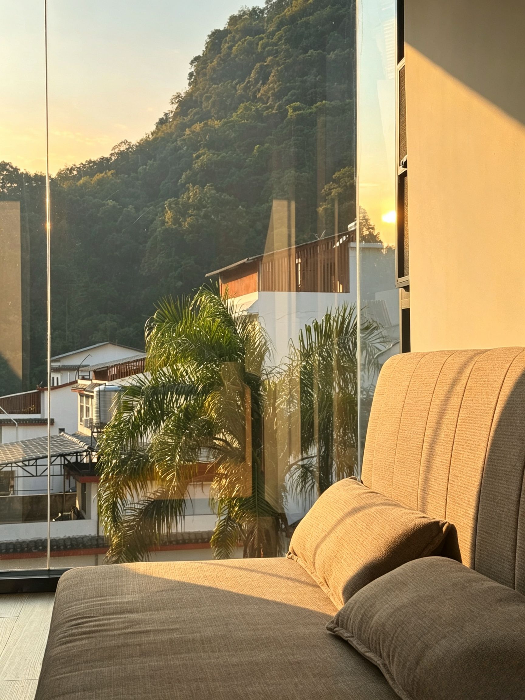 A room with a large window overlooking a hillside, a sofa, and a view of a building with a sloped roof.