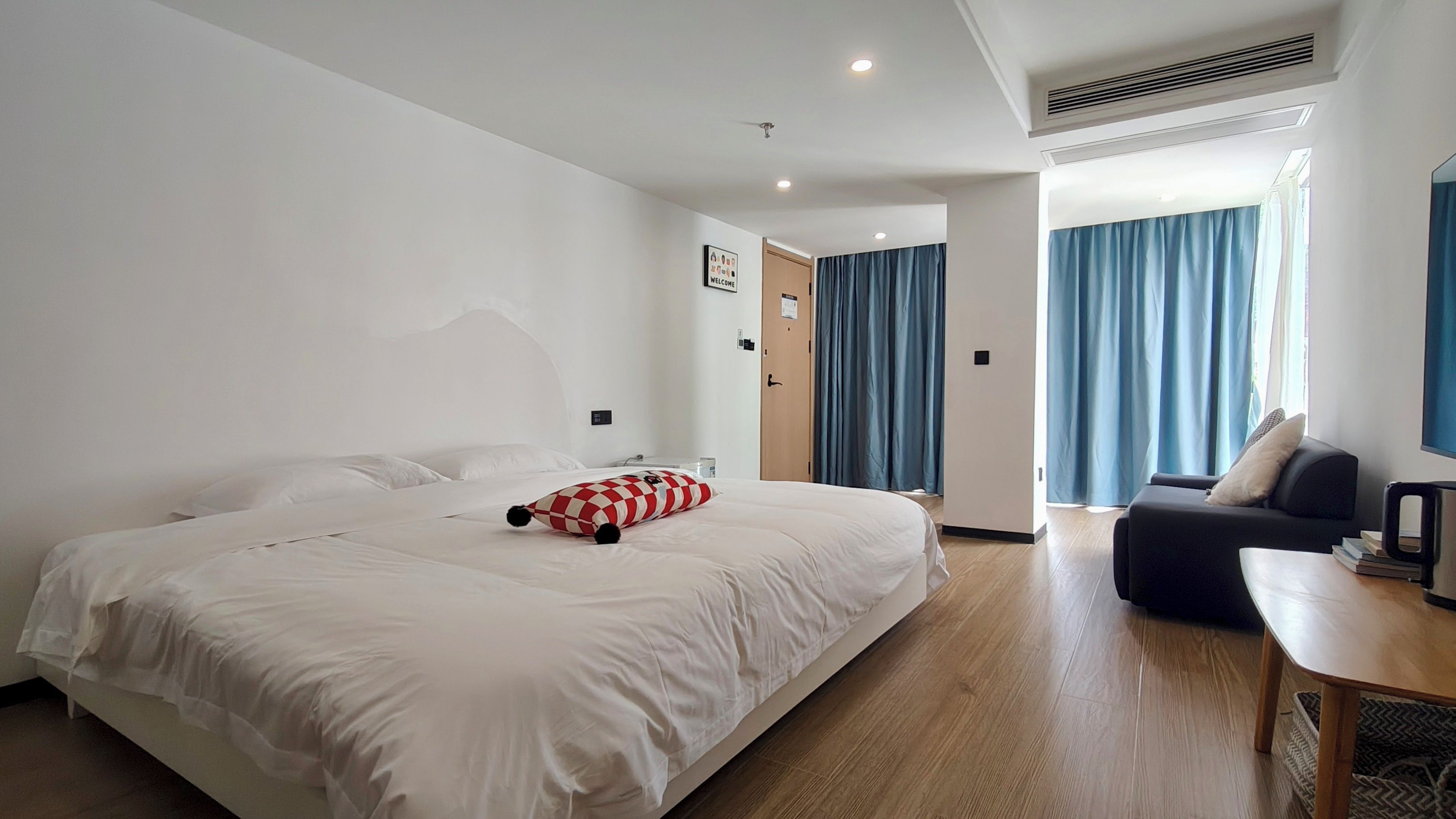 A hotel room with a large bed, a small sofa, a wooden coffee table, and a view of the outside through a window with blue curtains.