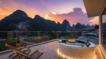 A balcony with a round bar area, lounge chairs, and a view of mountains at sunset.