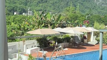 A poolside view with a mountain backdrop, a dining table with fruit and bread, and outdoor seating with umbrellas.