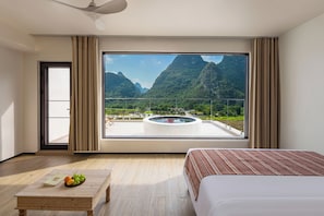 Habitación de hotel con un ventanal grande, una cama, una mesita con fruta y vistas a montañas.