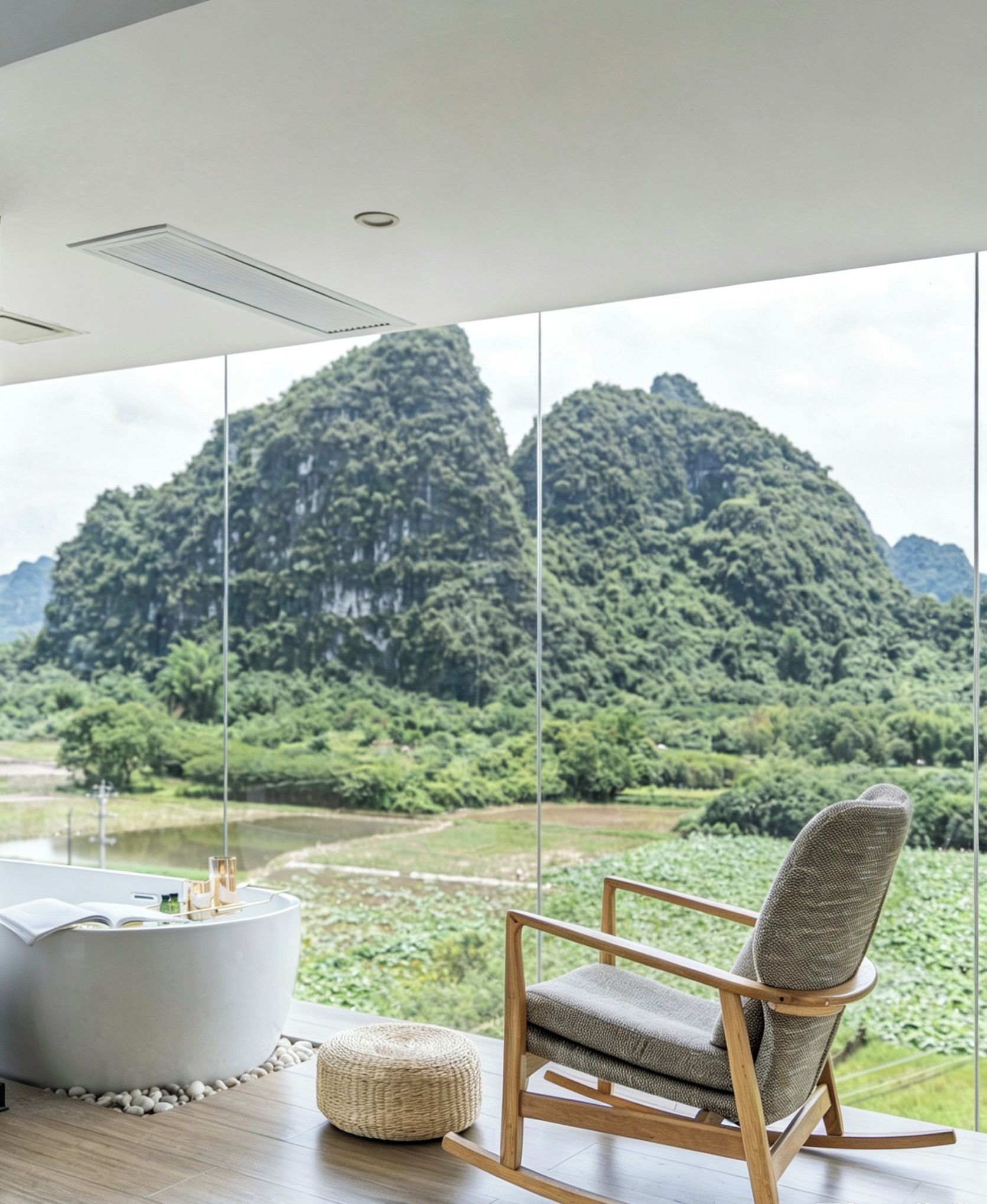 Habitación con vistas a un paisaje montañoso a través de amplias ventanas. Hay una mesa redonda blanca con un libro y una botella pequeña, y una mecedora de madera con un cojín.