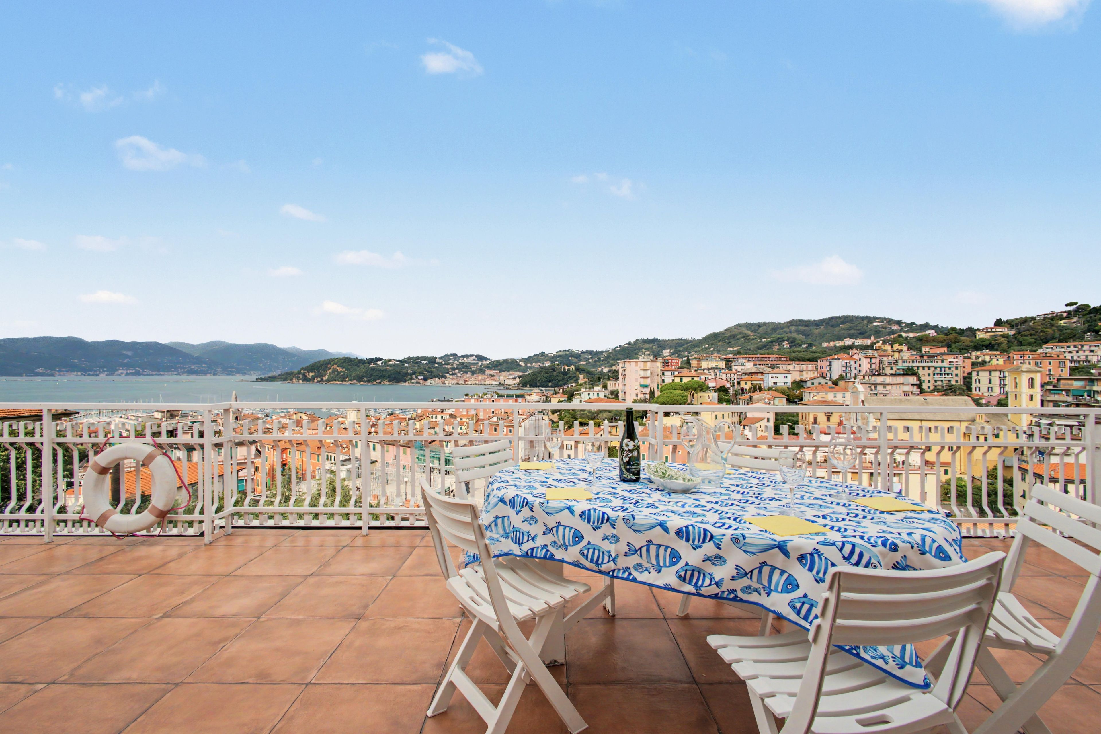 Una terrazza con un tavolo apparecchiato per due, con vista su una località costiera, edifici e colline sullo sfondo.