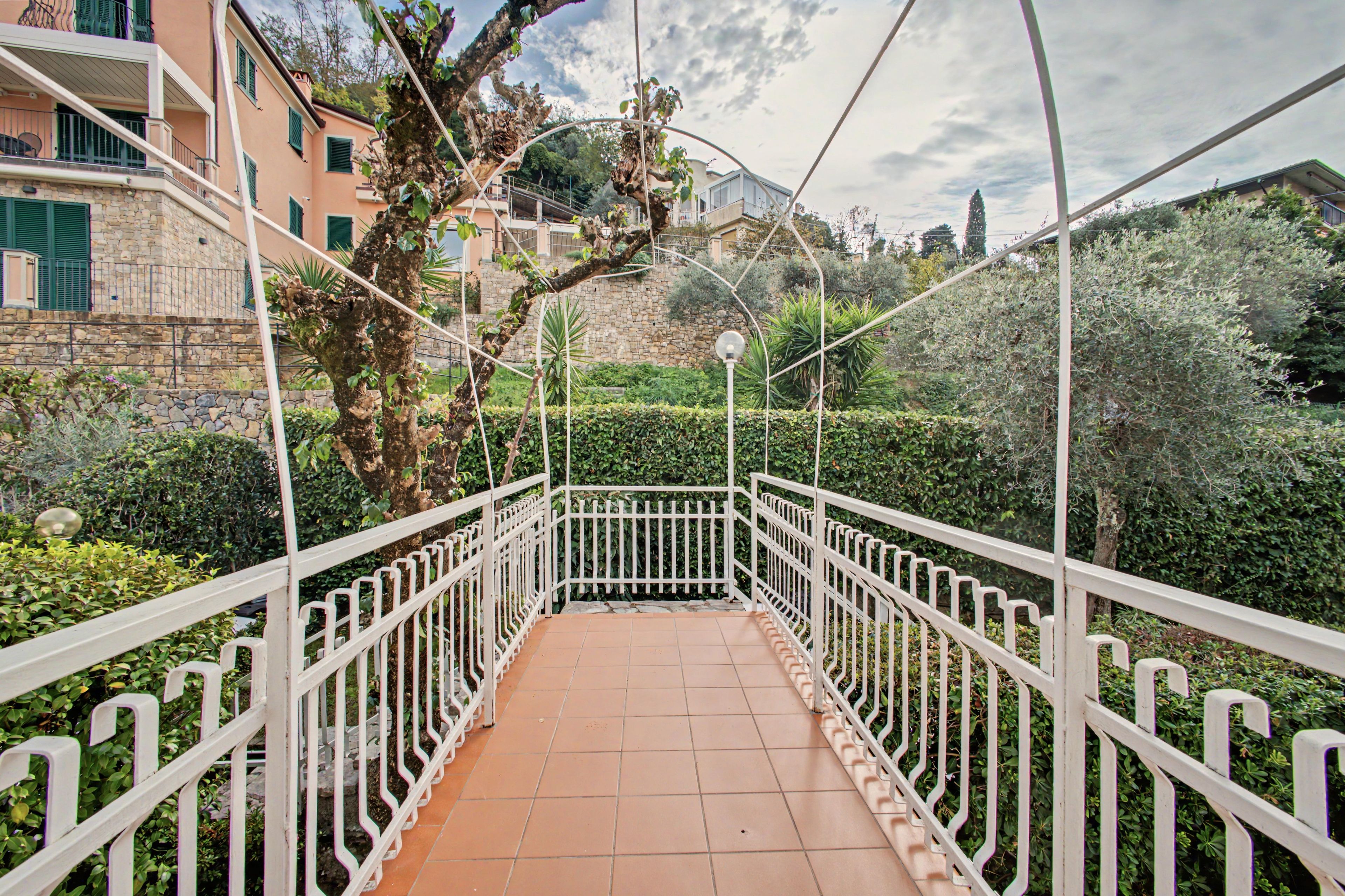 A pedestrian bridge with white railings, surrounded by greenery and buildings.