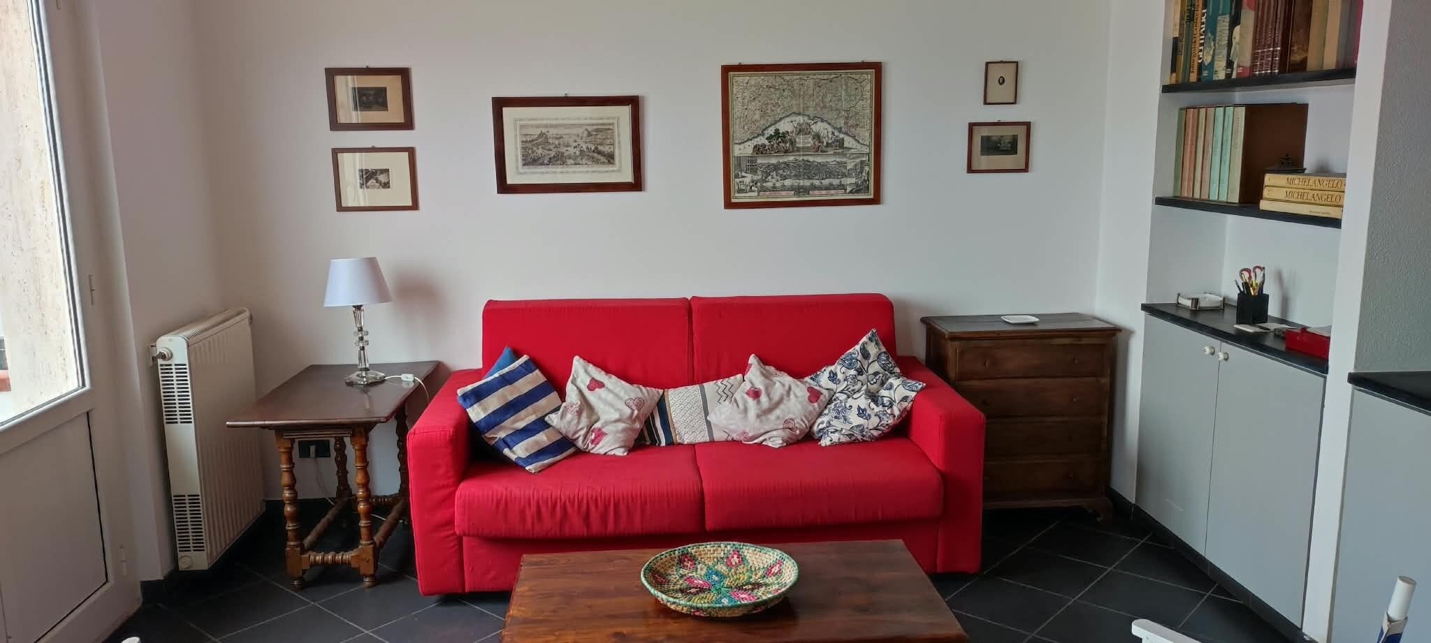 A red sofa with patterned cushions, a small wooden table, a radiator, and framed pictures on the wall.