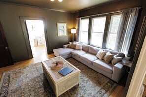 A living room with a sectional sofa, a coffee table, and a rug. There is a door leading to another room and a window with curtains. -  Spacious 4-Bedroom Group and Family Retreat: Game Room, Bunk Room, Walk2Shops (Matthews)