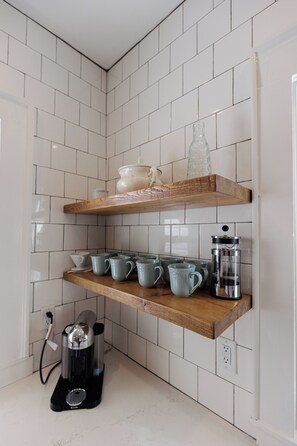 A kitchen with white tiled walls, wooden shelves, and various kitchen items including a coffee maker and cups. -  Spacious 4-Bedroom Group and Family Retreat: Game Room, Bunk Room, Walk2Shops (Matthews)