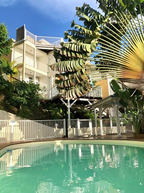 A pool area with a white fence, a multi-level building with balconies, and a large tropical plant. - Duplex Papaye Deshaies. Sea view. Swimming pool (Deshaies)