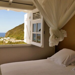 A bedroom with a bed, a window with curtains, and a view of a coastal landscape. - Copy of Duplex Papaye Deshaies. Sea view. Swimming pool (Deshaies)