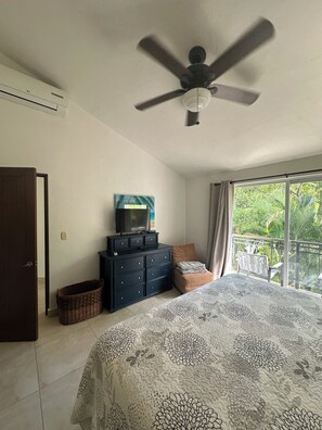 Una camera da letto con un letto, una cassettiera, una ventola a soffitto, una finestra vista su del verde e una porta.