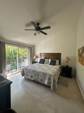 Une chambre à coucher comprenant un lit, un ventilateur de plafond, des tables de chevet et une vue sur un balcon accessible par une porte coulissante.
