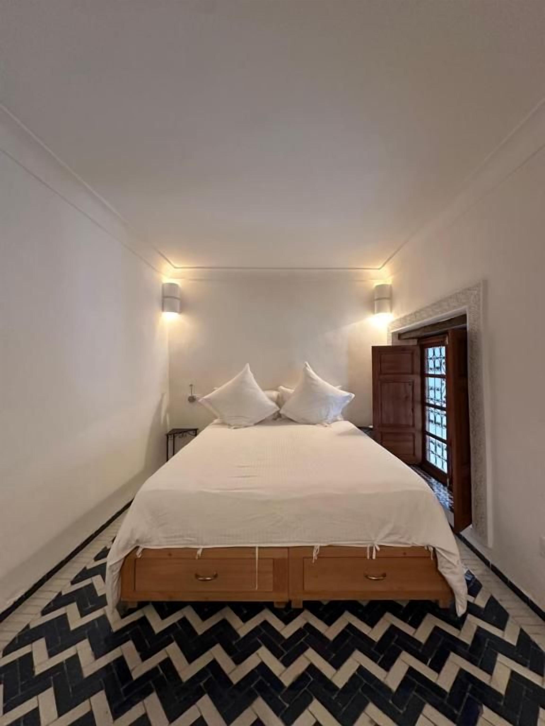 Una habitación pequeña con una cama, dos lámparas de pared, un banco de madera y una puerta con ventanal.