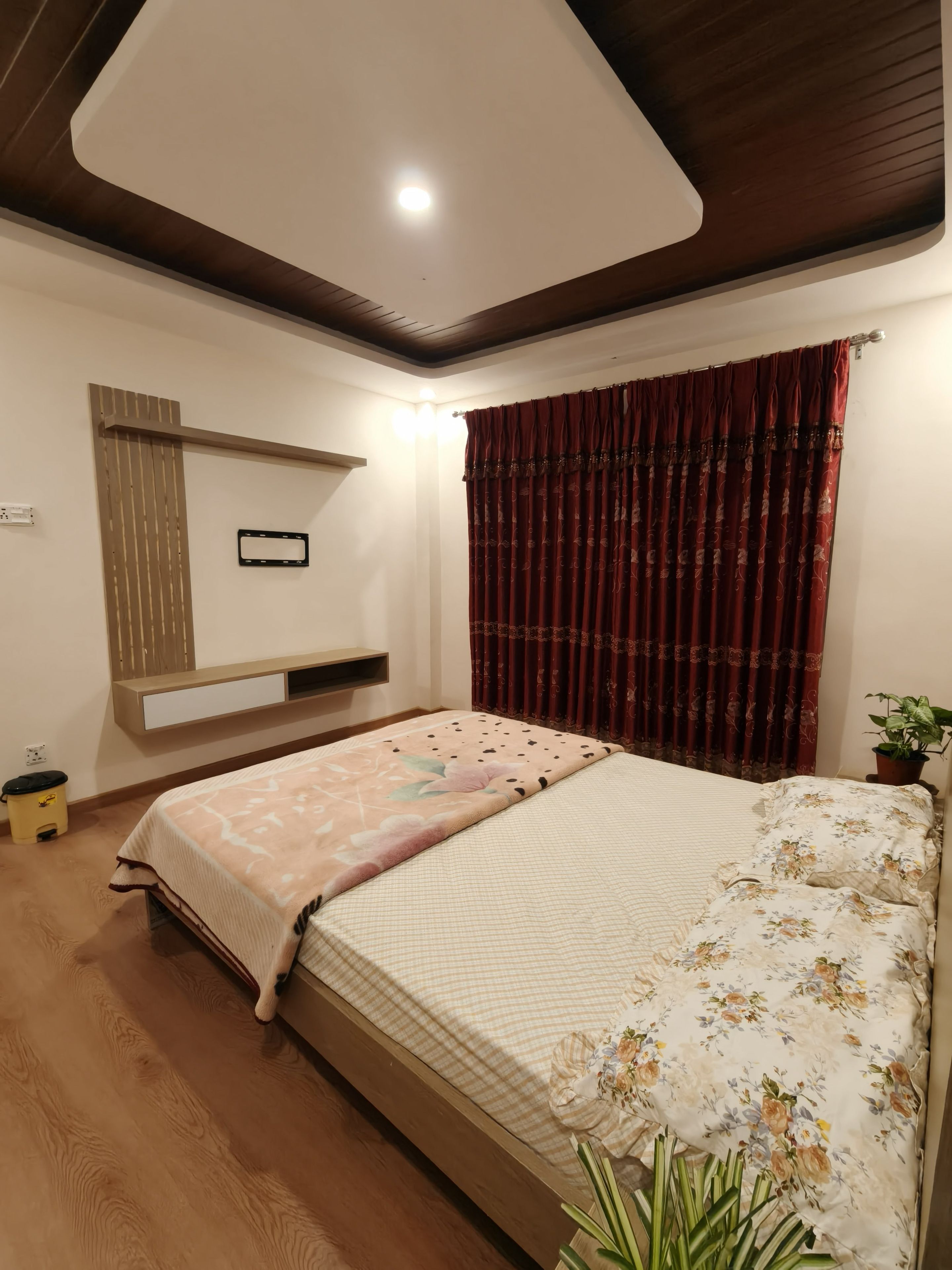 Un dormitorio con cama, mesita de noche, estante en la pared, planta en maceta y luz de techo.