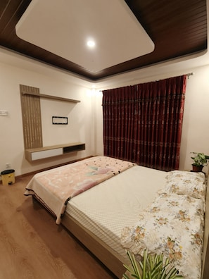 A bedroom with a bed, a bedside table, a wall-mounted shelf, a potted plant, and a ceiling light.