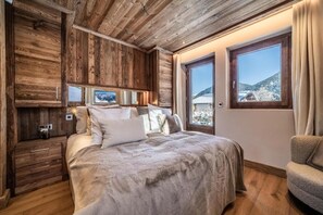 A bedroom with a wooden ceiling, a large bed with multiple pillows, a nightstand, and a view of a snowy landscape through a window. - Luxury Mountain Chalet with Jacuzzi, Sauna & Hammam near Mont Blanc (Saint-Gervais-les-Bains)