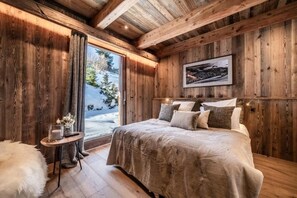 A cozy bedroom with a wooden ceiling, a large bed, a small bedside table, and a window with a view of trees. - Luxury Mountain Chalet with Jacuzzi, Sauna & Hammam near Mont Blanc (Saint-Gervais-les-Bains)