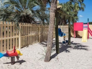 A sandy playground area with a wooden fence, palm trees, and a swing set. - Summer House 1006A (Orange Beach)