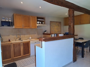 A kitchen with wooden cabinets, a built-in microwave, a sink, and a dining area with a table and chairs. - Holiday Home 'Longues Amarres' with Terrace, Garden and Wi-Fi (Longues-sur-Mer)