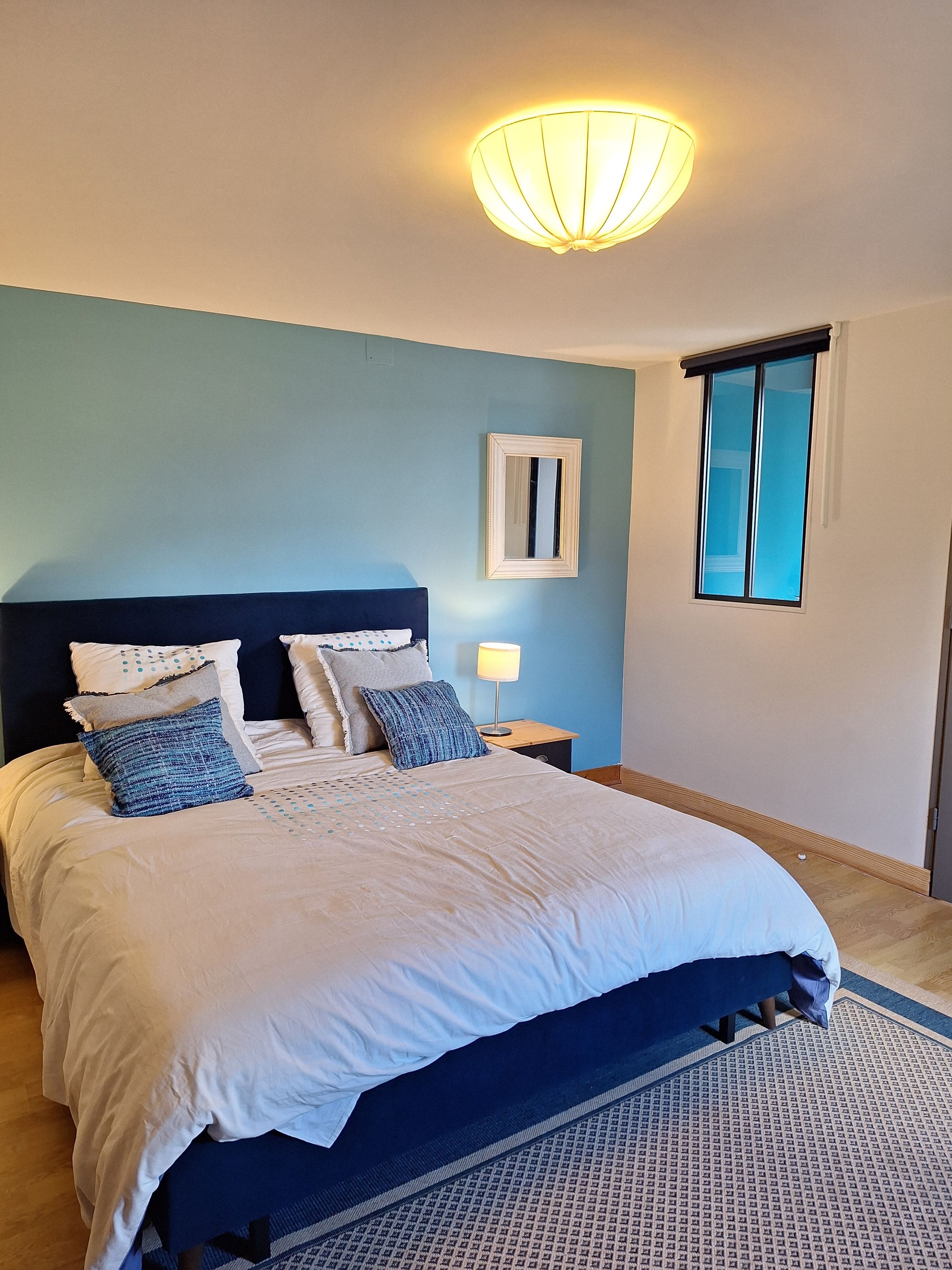 Une chambre à coucher comprenant un lit, une table de chevet, un miroir fixé au mur et une fenêtre avec des stores.