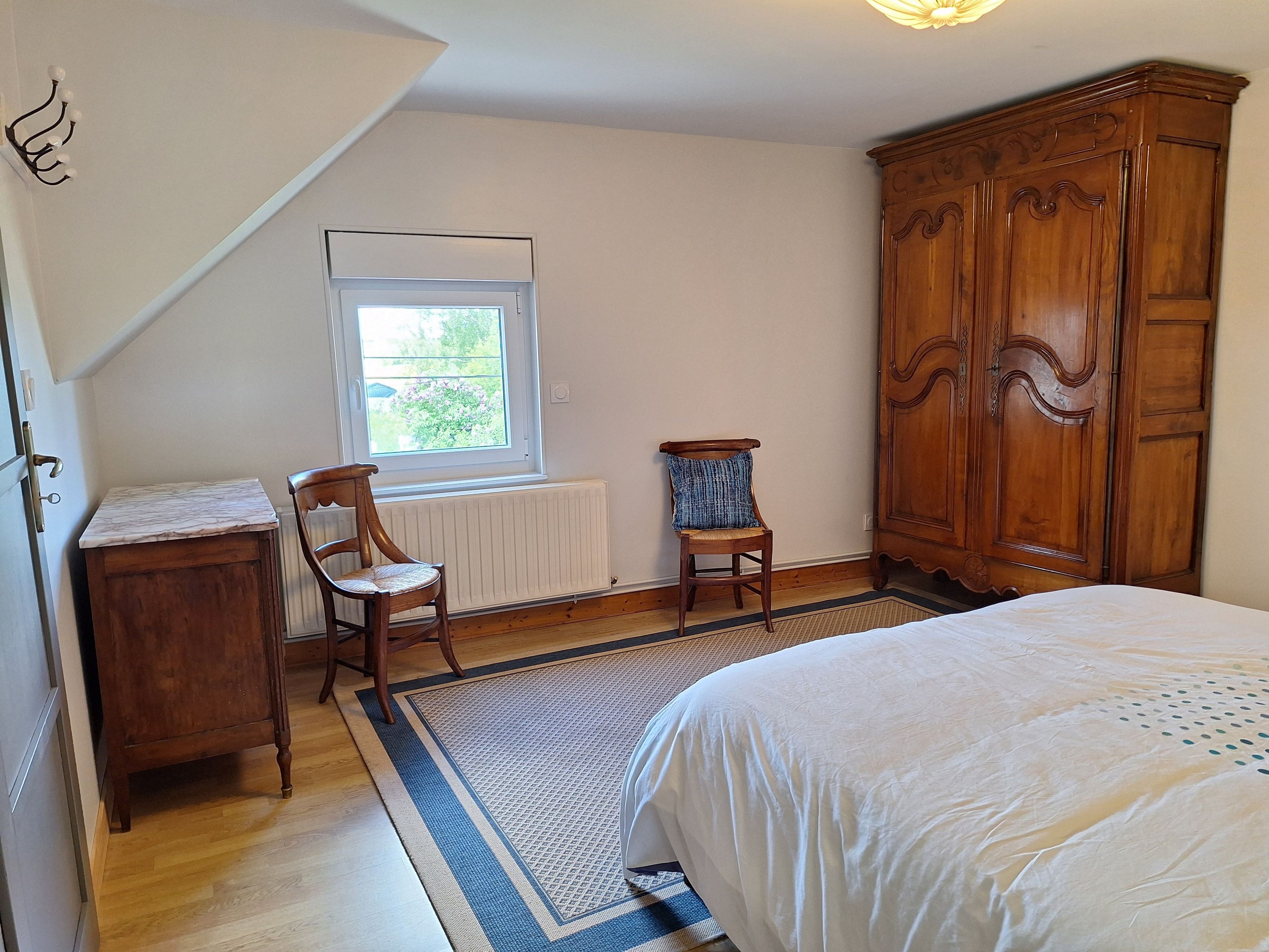 Een slaapkamer met een bed, een houten garderobe, een stoel, een radiator en een raam met uitzicht.