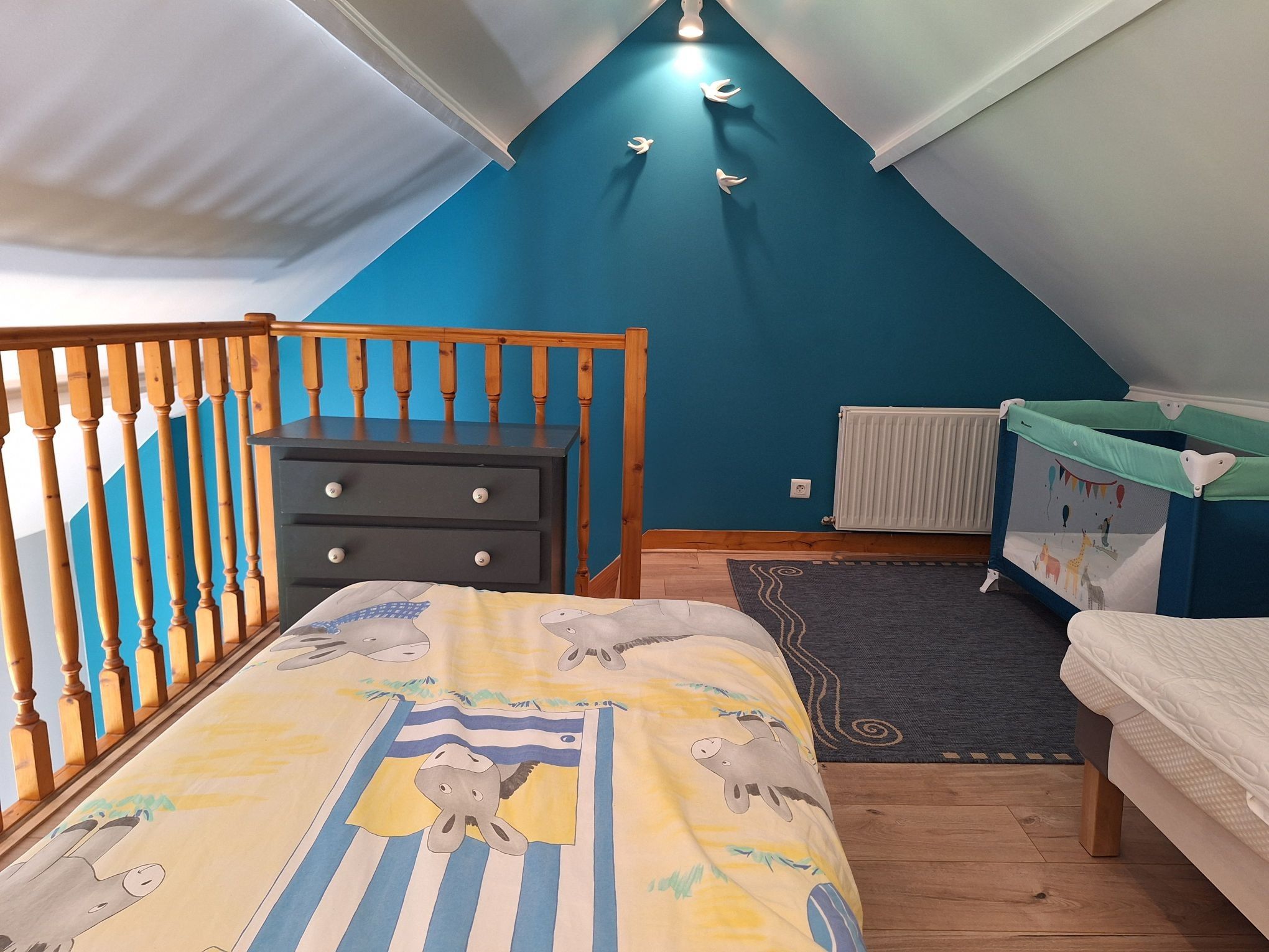 A bedroom with a sloped ceiling, a dresser, a bed with a dinosaur-themed bedspread, a crib with a colorful mural, and a radiator.