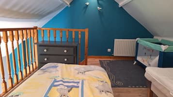 A bedroom with a sloped ceiling, a dresser, a bed with a dinosaur-themed bedspread, a crib with a colorful mural, and a radiator.