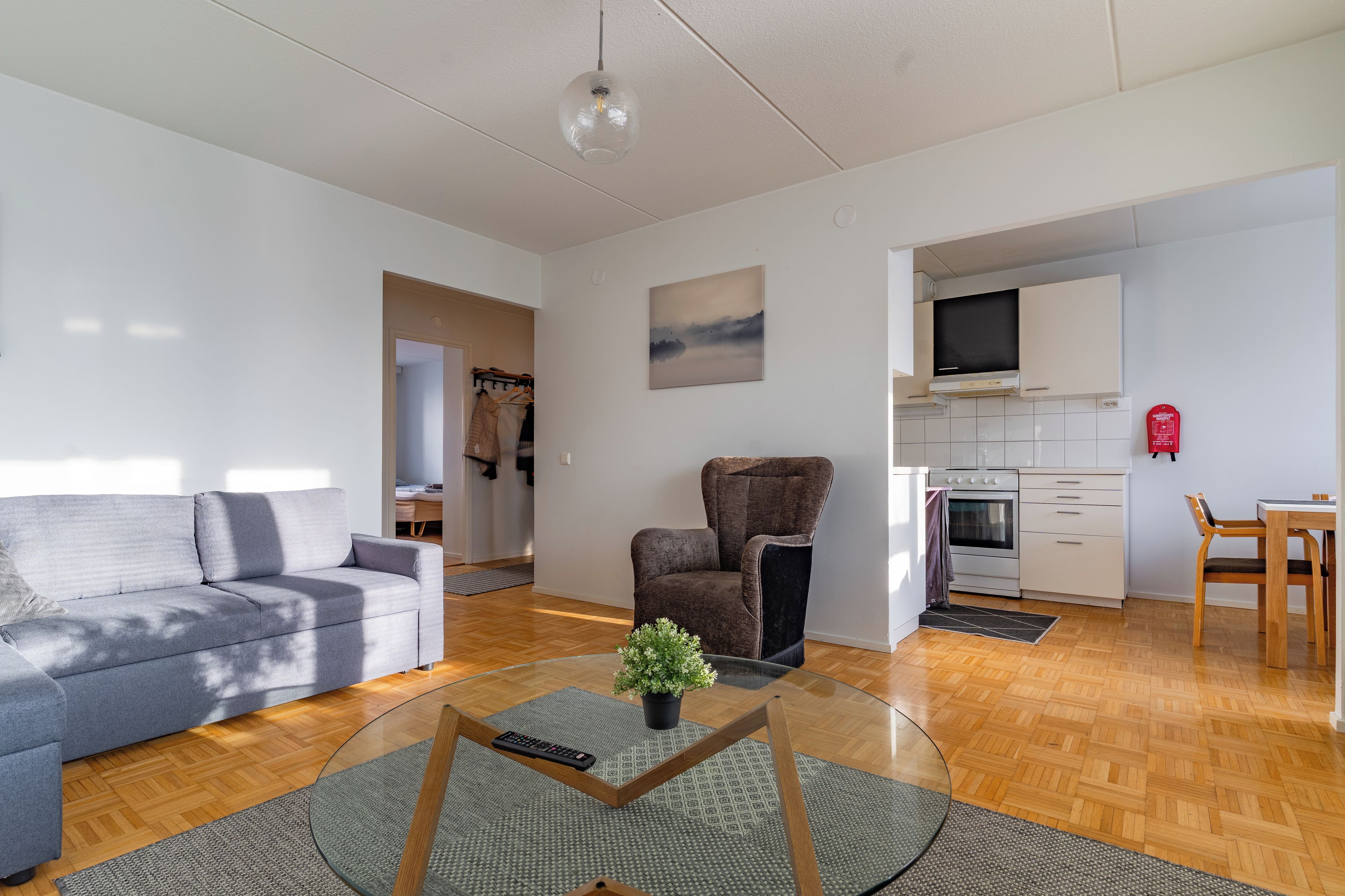 Een moderne woonkamer met een grijze bank, een bruine fauteuil, een glazen salontafel met een afstandsbediening, en een kleine plant.