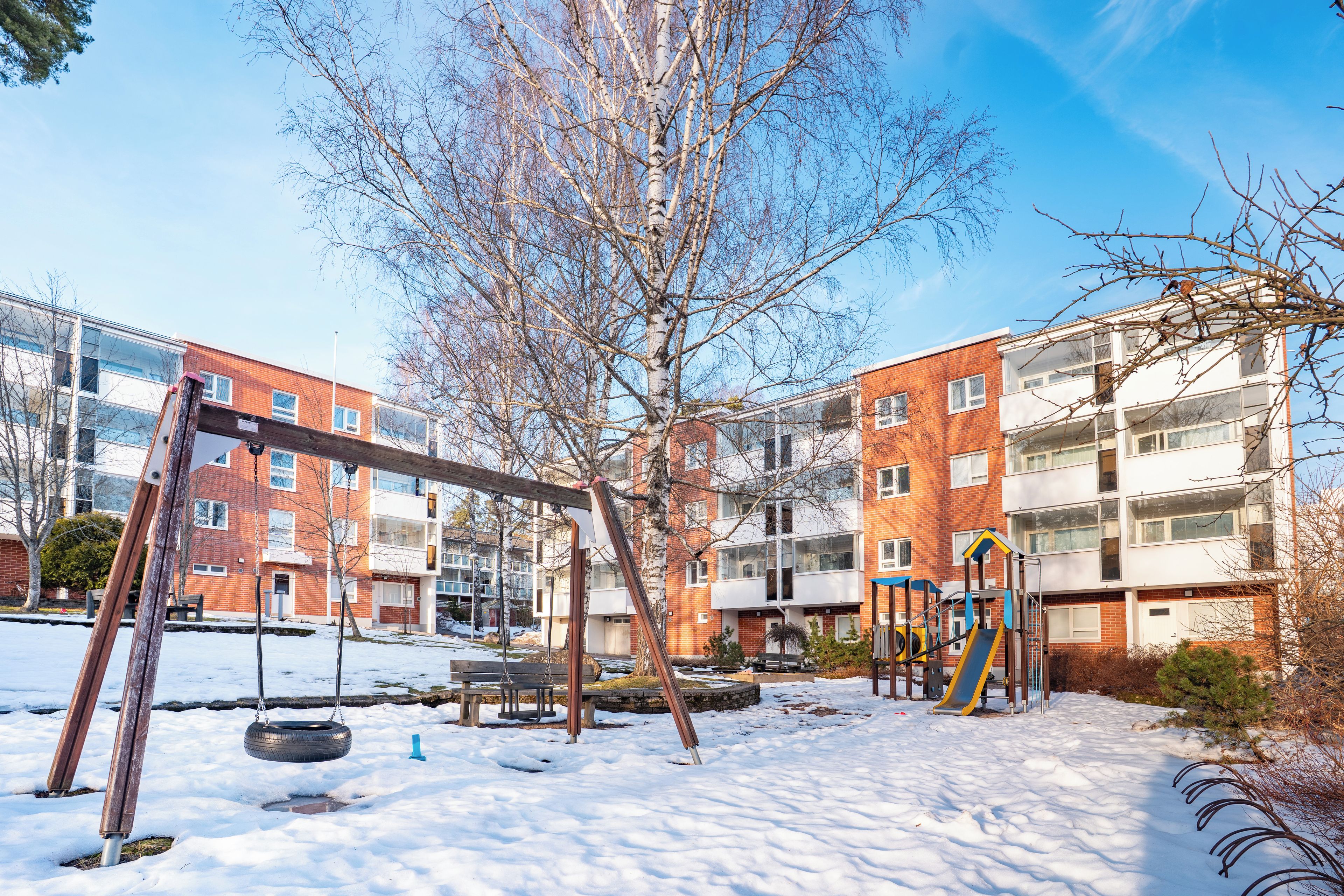 Un'area giochi con altalene e scivoli, circondata da terreno innevato e un edificio con numerose finestre.
